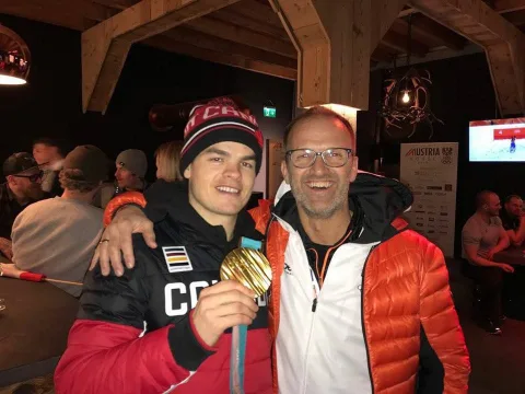 Ein Sportler mit Goldmedaille und ein Begleiter stehen Arm in Arm in einer Hütte