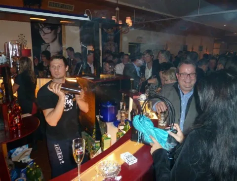 Eine Gruppe von Menschen hat sich in einer Bar versammelt, darunter ein Mann, der ein schwarzes T-Shirt mit der Aufschrift „Just do it“ trägt.