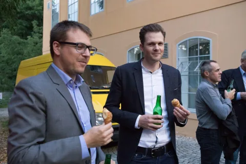 Zwei Männer in Anzügen stehen nebeneinander und halten Bierflaschen in den Händen.