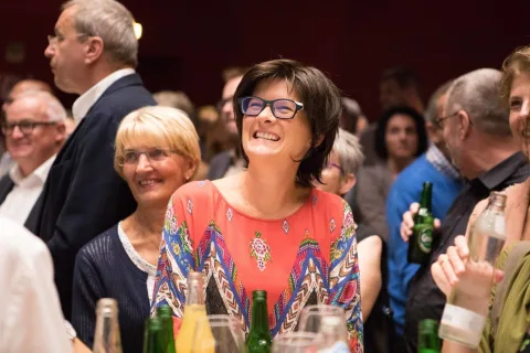 Eine Frau mit Brille lacht in einer Menschenmenge auf einer Party.