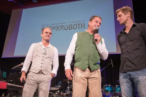 Drei Männer stehen nebeneinander auf einer Bühne und einer von ihnen hält ein Mikrofon.