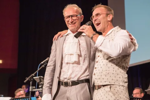 Zwei Männer stehen nebeneinander auf einer Bühne und einer singt in ein Mikrofon.