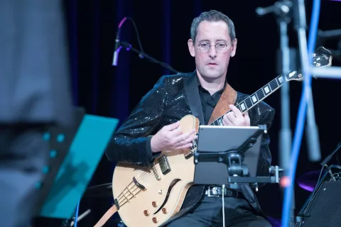 Ein Mann in einer Paillettenjacke spielt auf einer Bühne Gitarre.