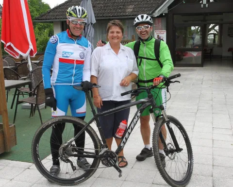 Zwei Radfahrer und eine Frau im weißen Polo stehen mit Rädern vor einem Gebäude