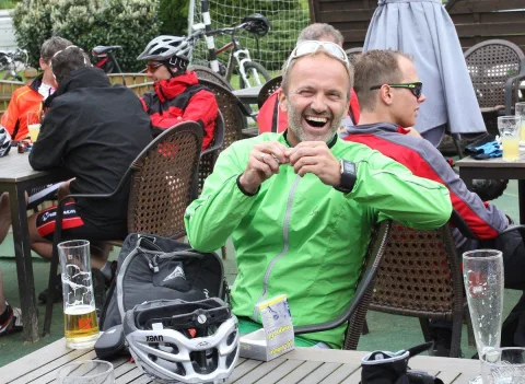 Radfahrer sitzen nach der Ausfahrt mit Bier an Tischen im Freien, einer lacht in die Kamera
