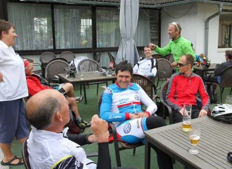 Radfahrer sitzen bei einer Rast an Tischen vor einer Hütte, einer prostet mit einem Bier