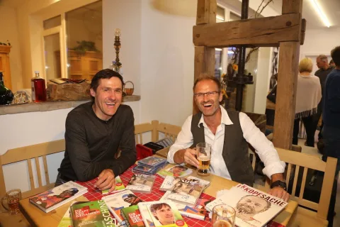 Zwei Männer sitzen an einem Tisch in einer Stube, vor ihnen liegen Zeitschriften