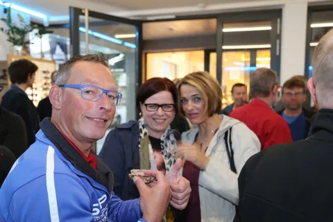 Drei Gäste stoßen mit Gläsern an, im Hintergrund das Schaufenster