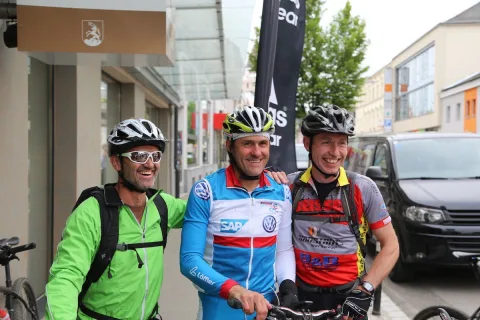 Drei Radfahrer stehen lachend nebeneinander, einer legt dem anderen den Arm um die Schulter