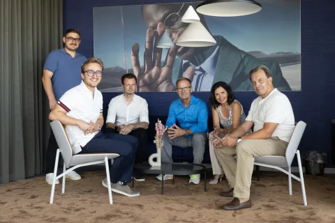 Sechs Personen sitzen und stehen vor einer dunkelblauen Wand mit einem großformatigen Foto, darüber kegelförmige Leuchten