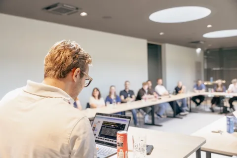 Ein Mann sitzt mit dem Rücken zur Kamera an einem Laptop, vor ihm die besetzten Tischreihen eines Saals